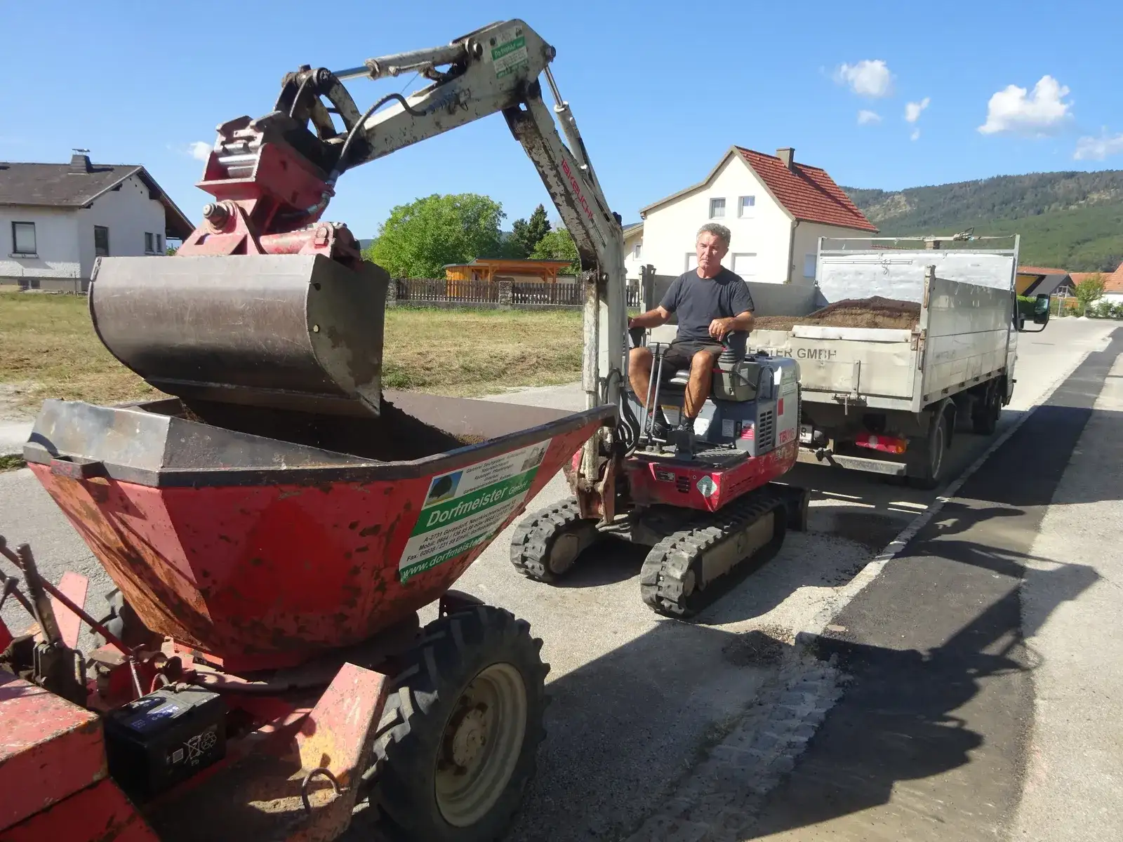 Minibagger belädt einen Dumper mit Erde, dahinter ein Lieferwagen in einer Wohnstraße