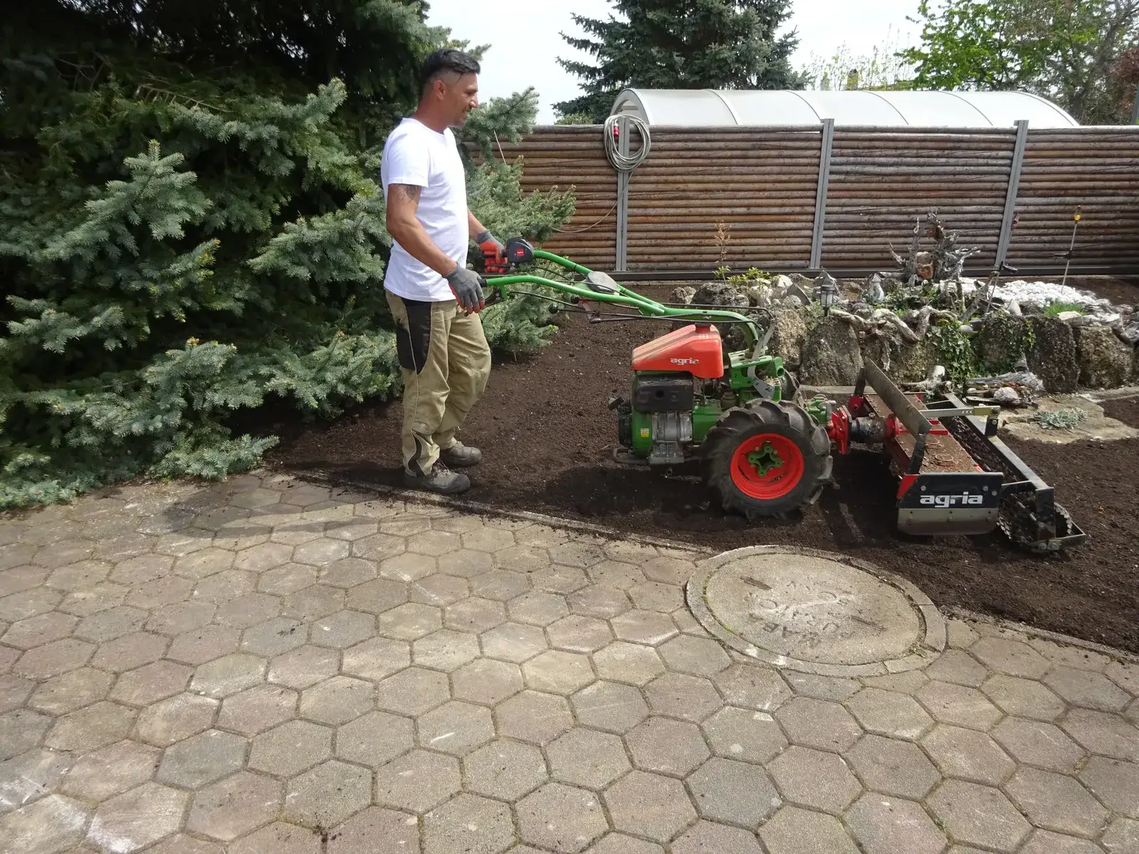 Mitarbeiter führt eine Einachs-Kreiselegge über dunkle Gartenerde neben einer gepflasterten Fläche