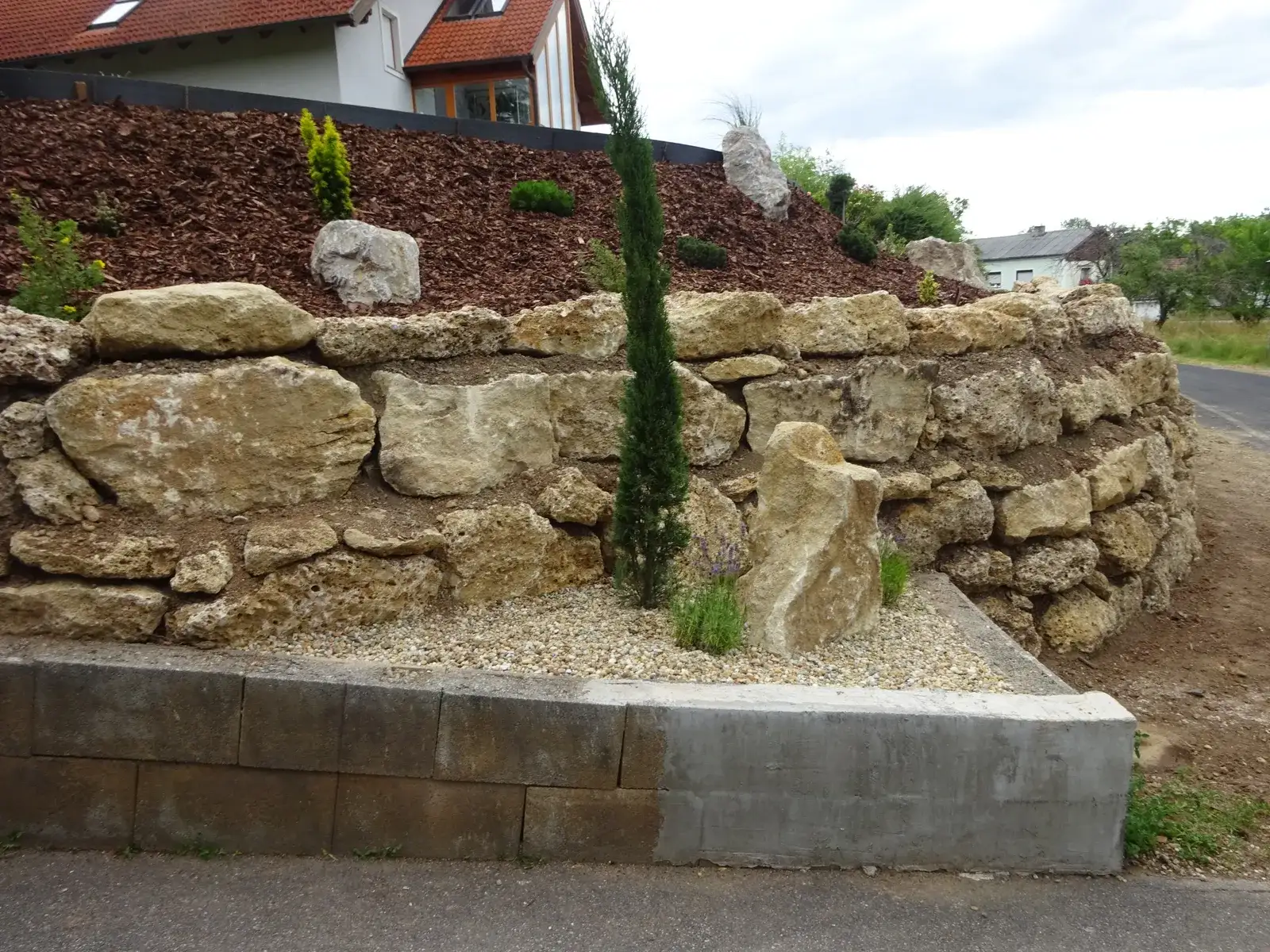 Fertige Natursteinmauer mit Kiesstreifen, Zypresse und Bepflanzung an einer Straße