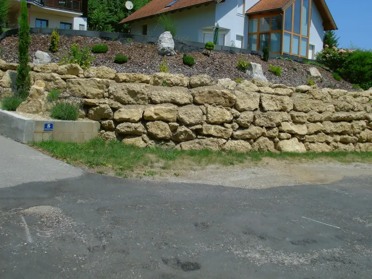 Fertige Natursteinmauer als Stützmauer vor einem Wohnhaus, dahinter der bepflanzte Hang