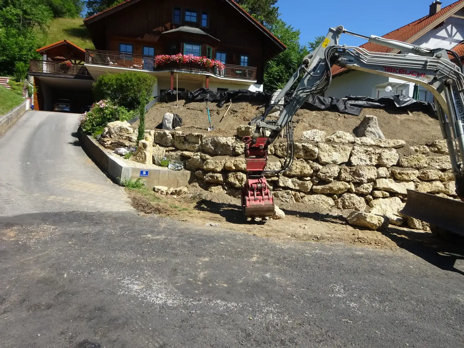 Bagger versetzt große Natursteine für eine Mauer entlang einer Hauszufahrt