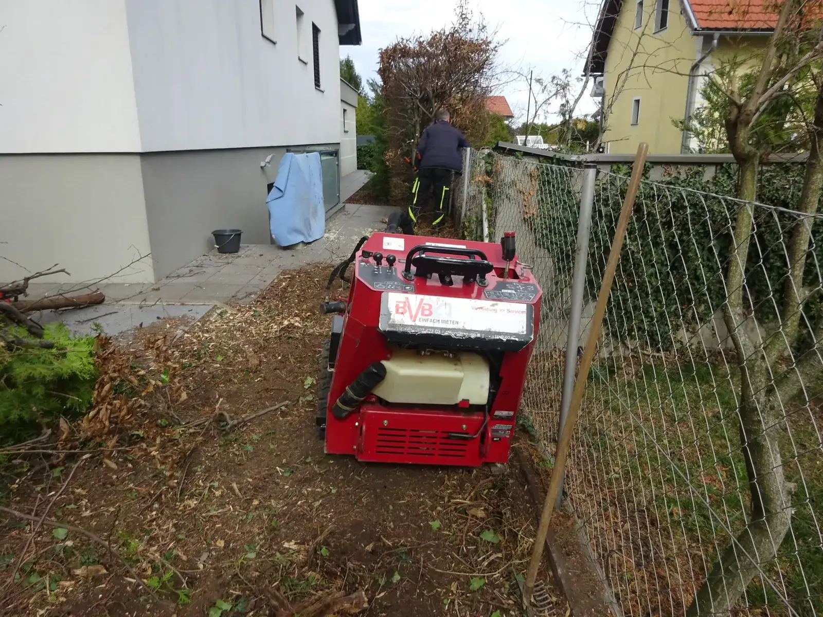 Stubbenfräse neben einem Maschendrahtzaun zwischen Haus und Garten